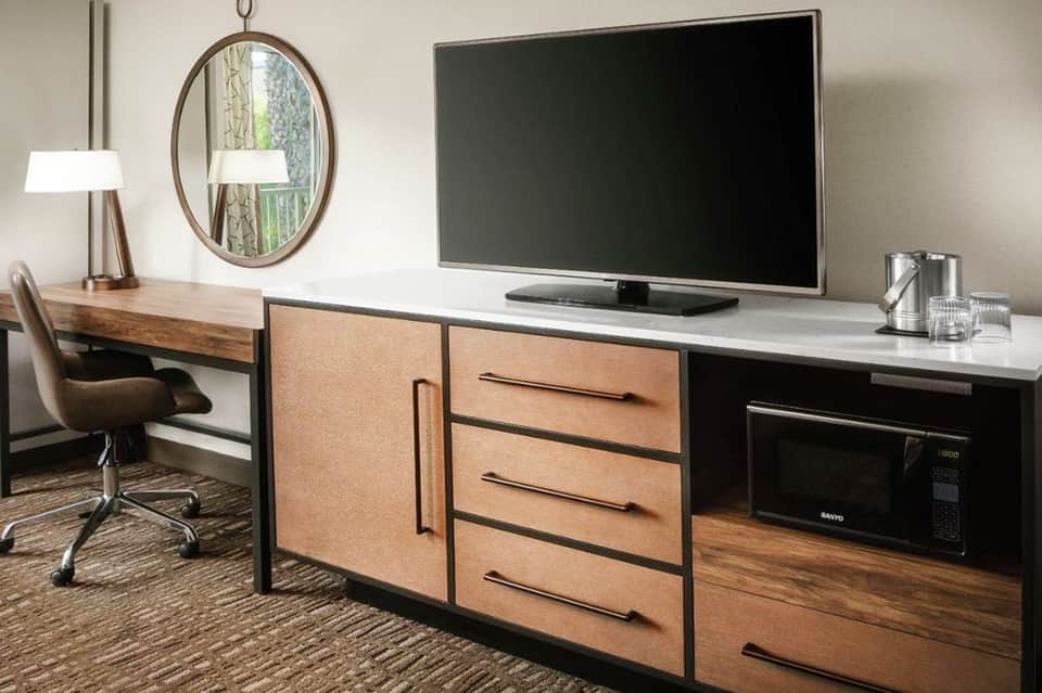 Modern hotel room desk with TV, wooden desk with drawers, desk lamp, and chair with oval mirror on wall