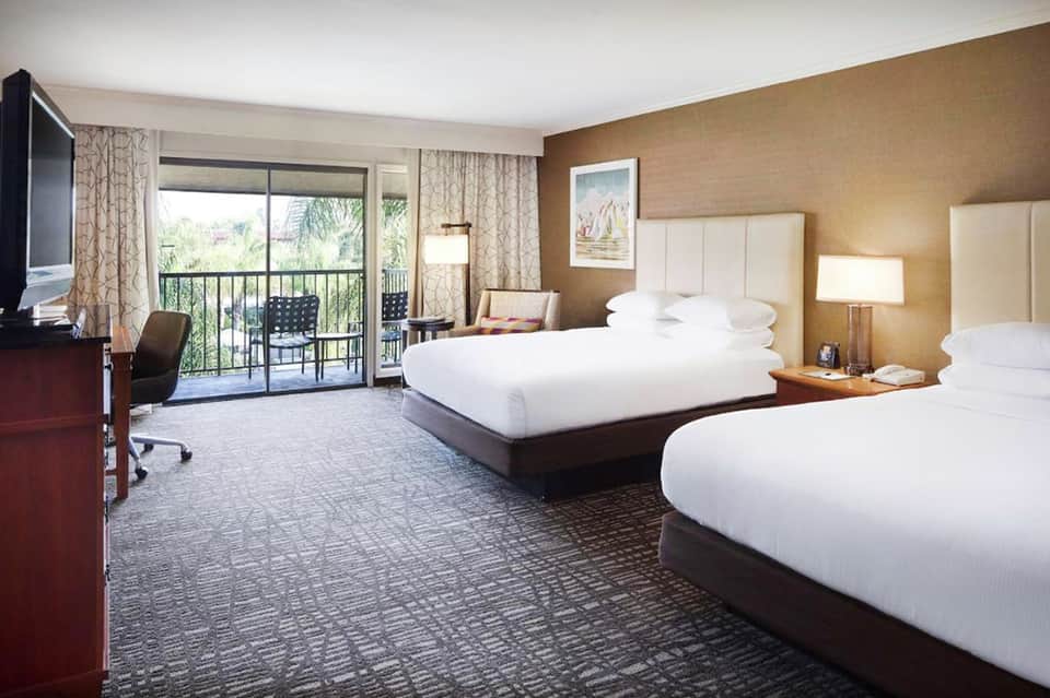 Hotel guest room with two queen beds, balcony access, flat-screen TV, and warm tan walls