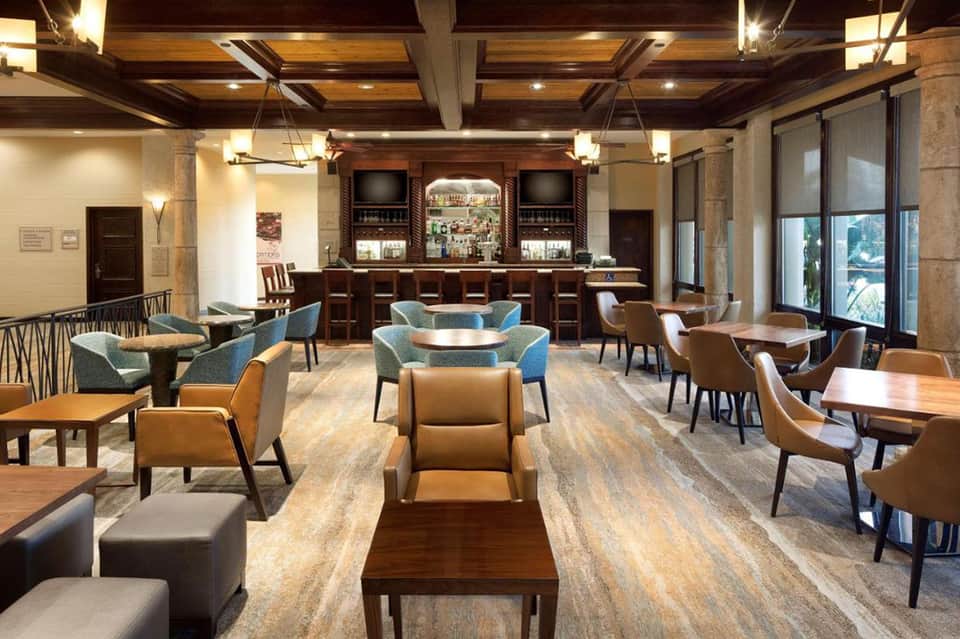 Upscale hotel lounge with wooden bar, leather seating, coffered ceiling, and large windows overlooking outdoor area