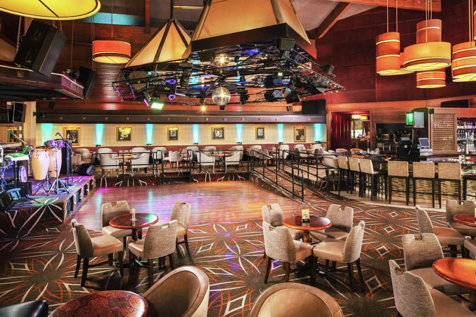 Modern nightclub lounge with colorful pendant lights, wooden bar, patterned carpeting, and stage setup