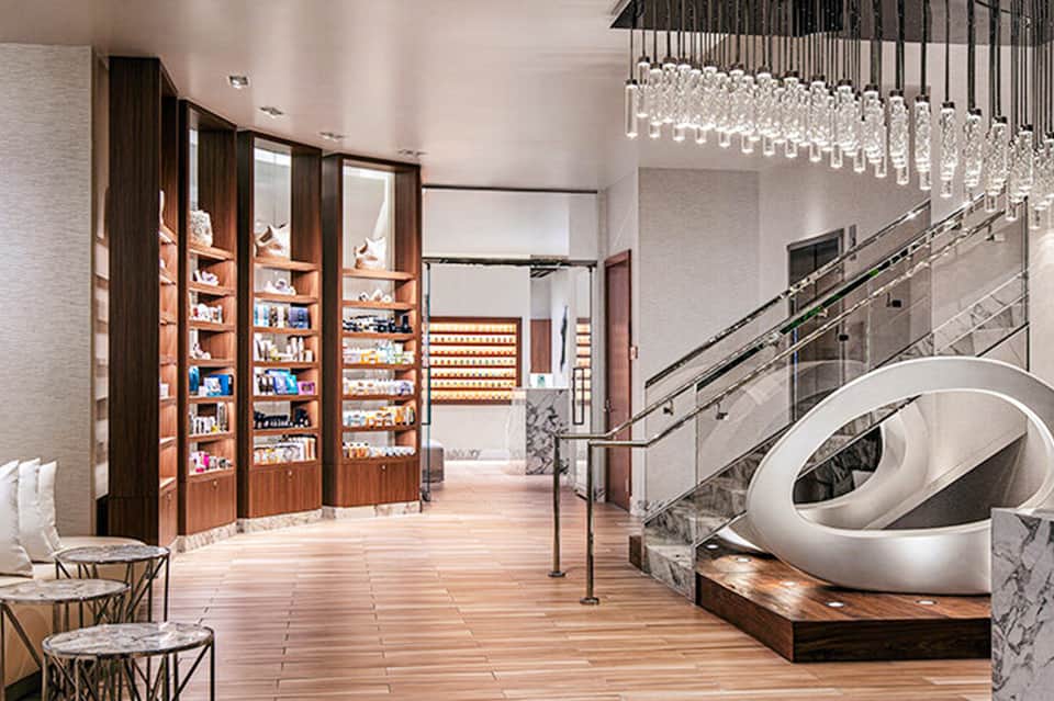 Modern luxury spa reception area with illuminated shelving, marble counters, and contemporary staircase with hanging glass chandelier