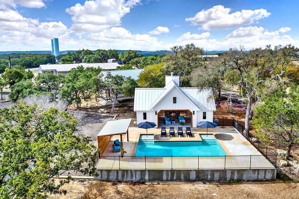 Modern white farmhouse with pool and lounge chairs surrounded by oak trees in Hill Country landscape