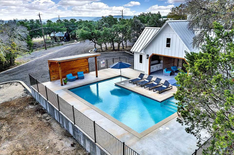 Modern poolside resort with turquoise lap pool, white cabana, lounge chairs, and surrounding oak trees on rural property