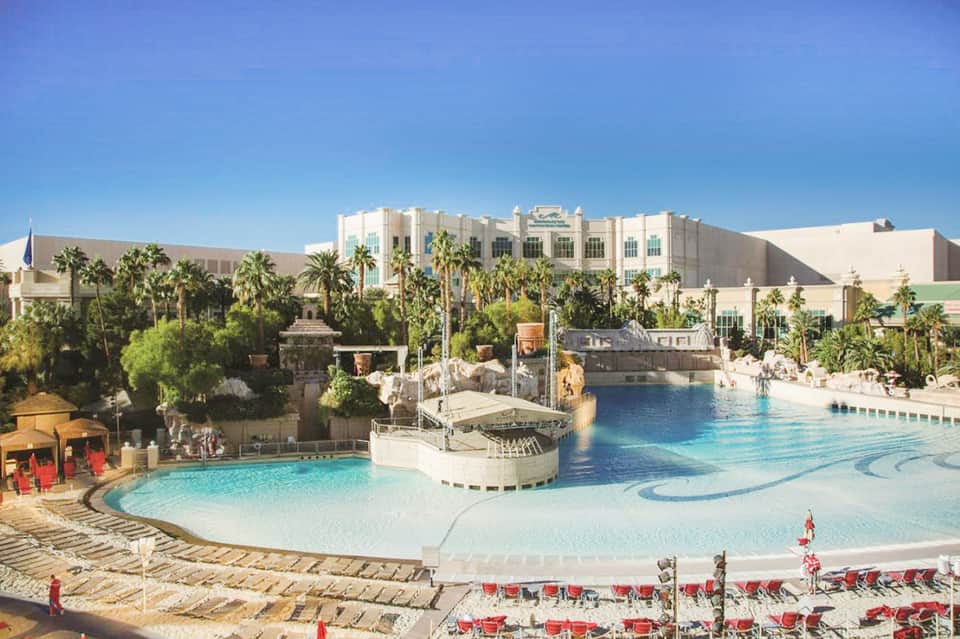 Large resort pool with turquoise water, palm trees, and white hotel building under clear blue sky