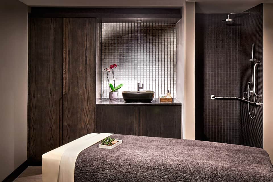 Spa treatment room with massage bed, dark wood accents, and ensuite bathroom with rainfall shower and window seating