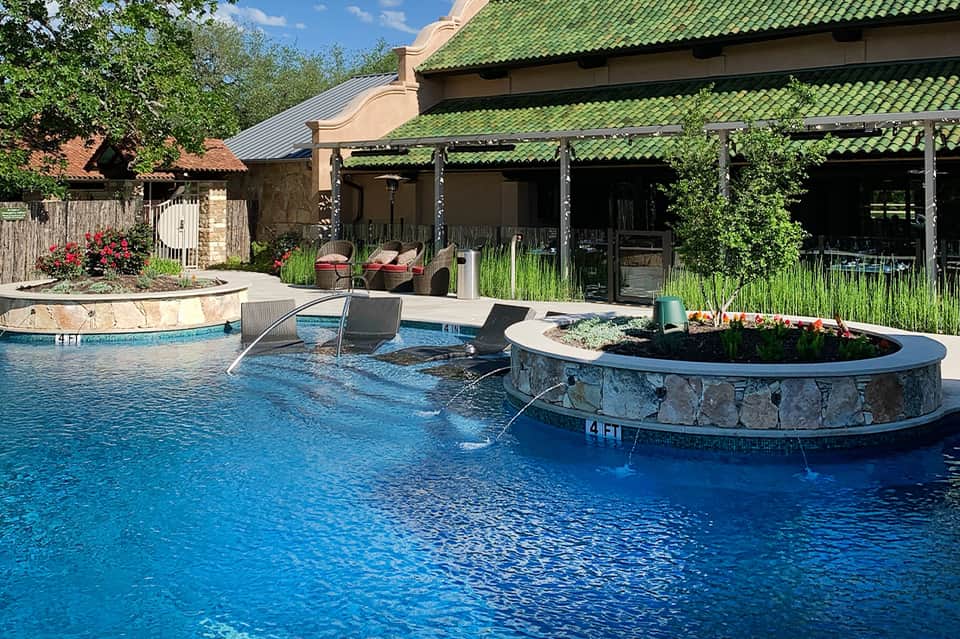 Resort pool with circular fire pit island, lounge chairs, and green-roofed building with pergola