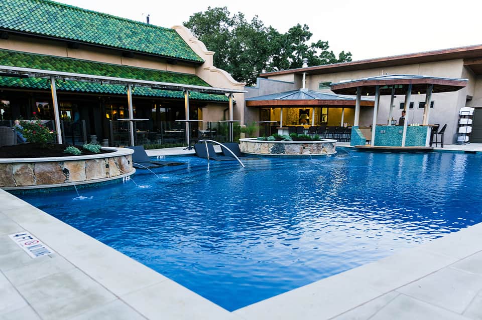 Resort pool with blue water, lounge chairs, green-roofed pavilion, and stone water features surrounded by buildings