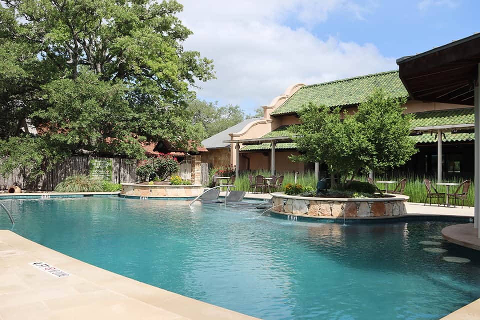 Resort courtyard with turquoise pool, spa area, stone fireplace, and green-roofed buildings surrounded by trees