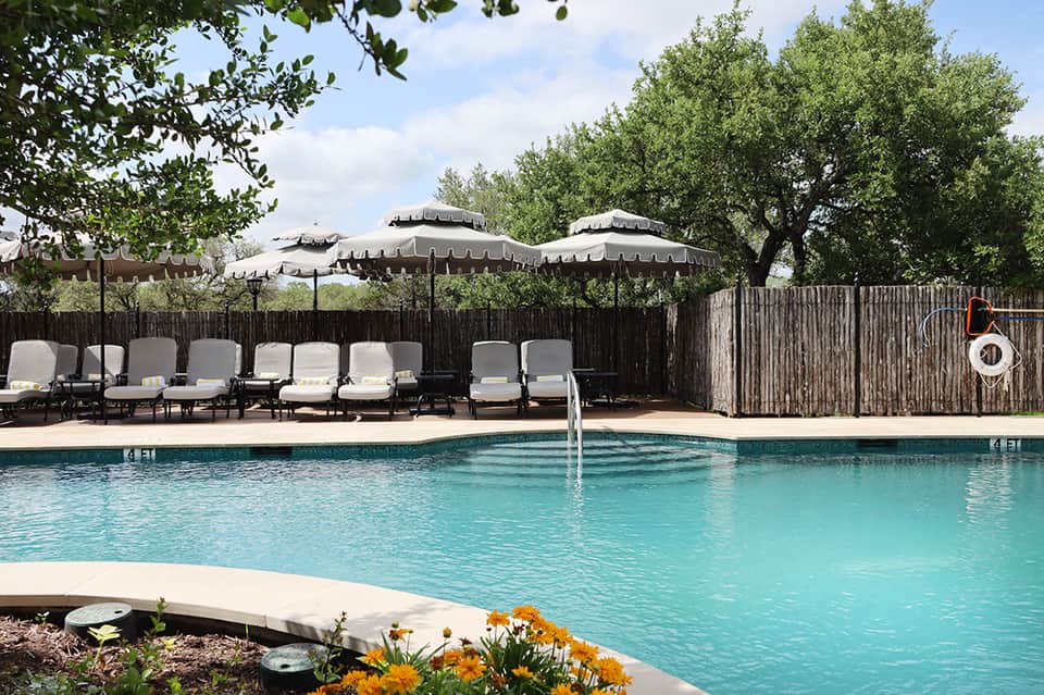 Turquoise resort pool with lounge chairs and umbrellas, surrounded by wooden fence and lush green trees