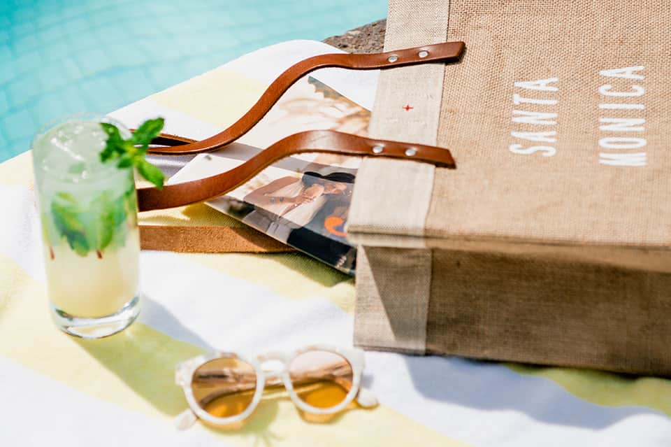 Poolside flat lay with mojito cocktail, sunglasses, beach bag, and leather sandals on white towel