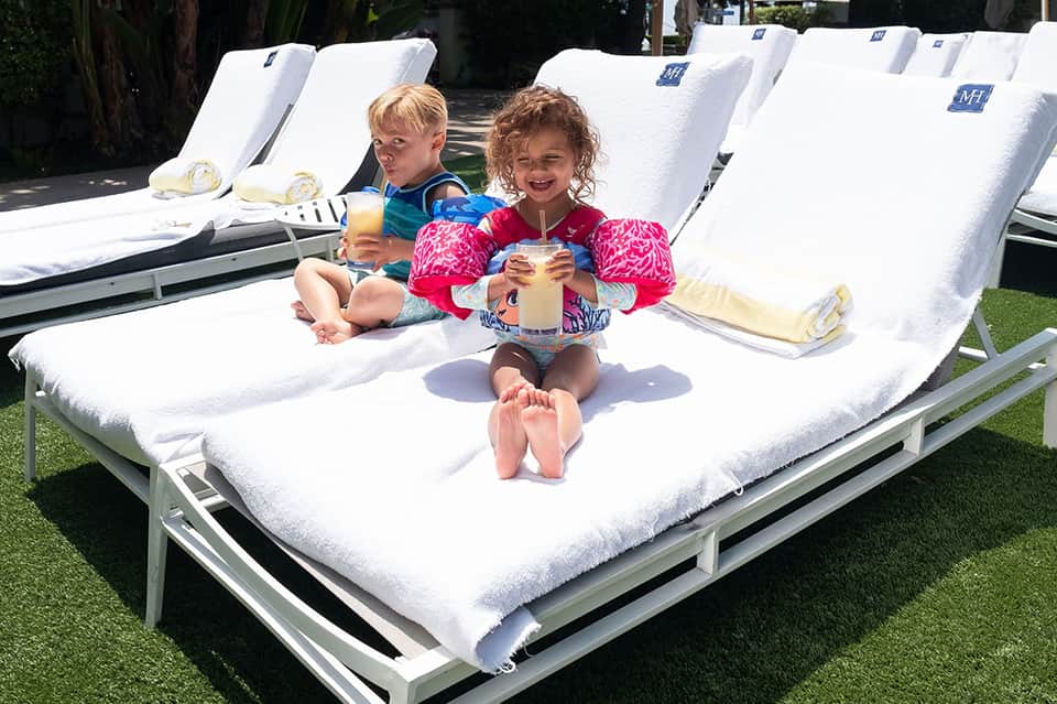 Two young children sitting on white lounge chairs by the pool with juice drinks