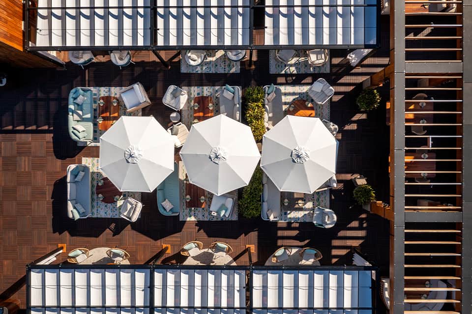 Aerial view of rooftop terrace with three white umbrellas, lounge seating, and dining tables on wooden deck