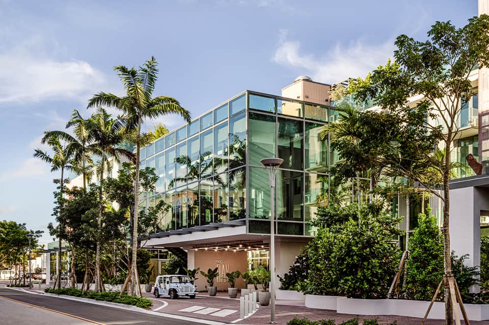 Modern glass-walled building with green-tinted windows surrounded by palm trees and tropical landscaping on a street corner.
