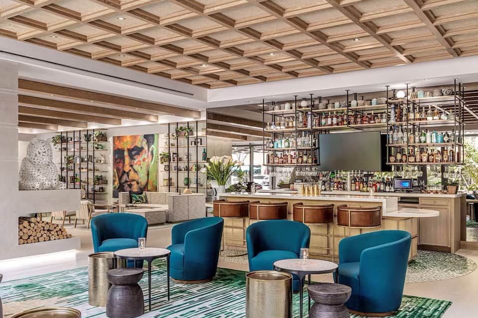 Modern hotel lounge with coffered wood ceiling, teal armchairs, and full-service bar with extensive bottle selection