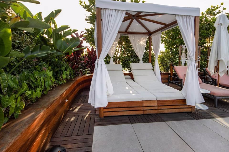 Luxury outdoor cabana bed with white curtains on wooden deck surrounded by tropical plants