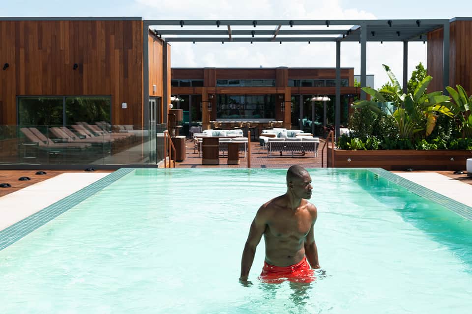 Man wading in turquoise pool with wooden modern building and pergola structure in background