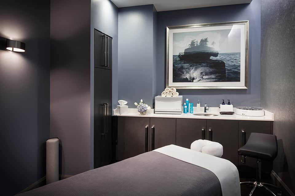 Modern spa treatment room with dark cabinetry, white massage bed, and ocean landscape artwork on blue wall
