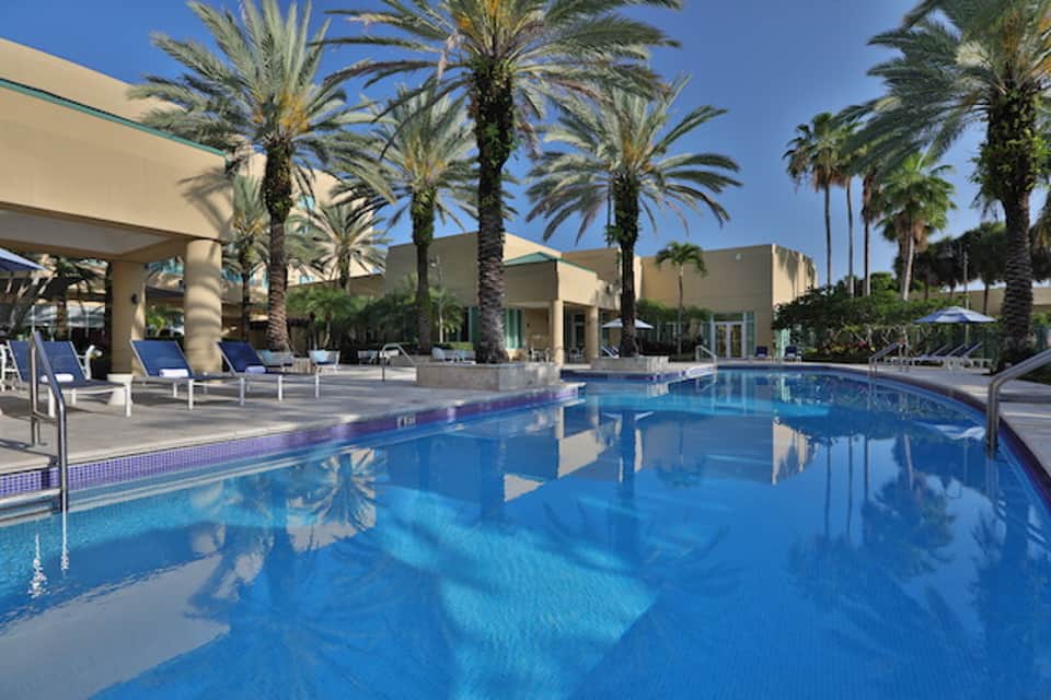 Resort pool with multiple lounge chairs, palm trees, modern pavilion structure, and clear blue water