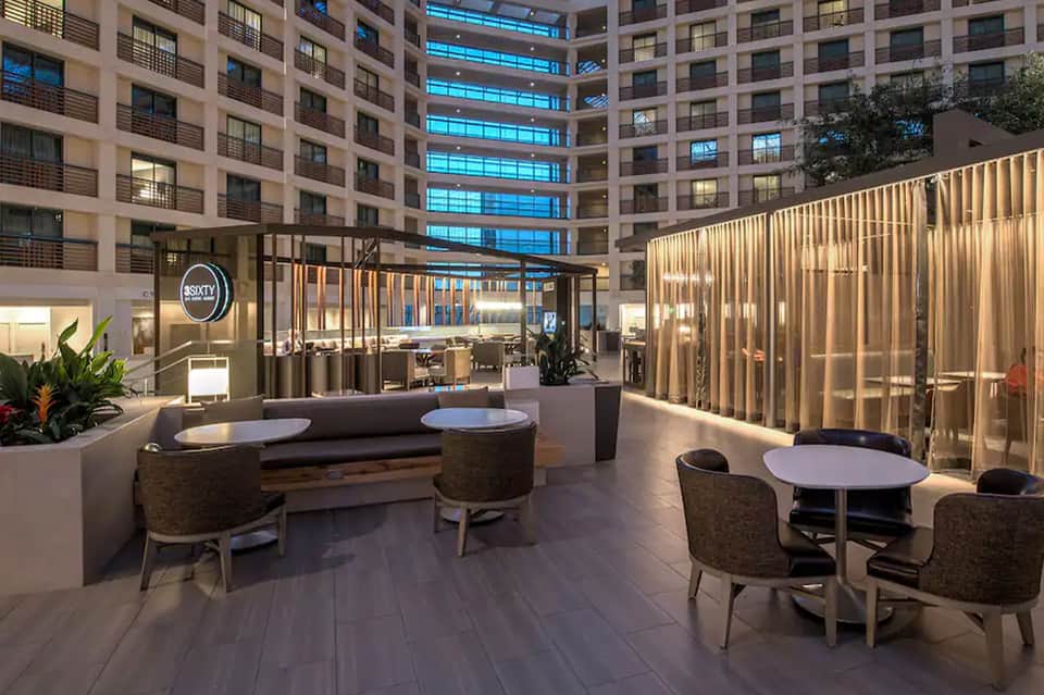 Modern hotel courtyard with lounge seating, round tables, and illuminated glass partitions under residential towers