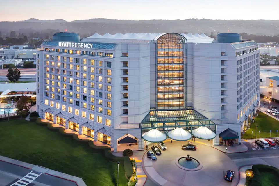 Hyatt Regency hotel at dusk with illuminated rooms, white tent structures in courtyard, mountains in background