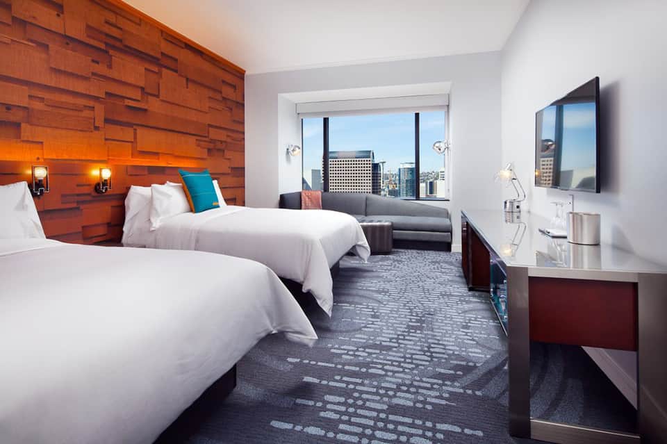 Modern hotel bedroom with two queen beds, wooden accent wall, city skyline views, and contemporary furnishings