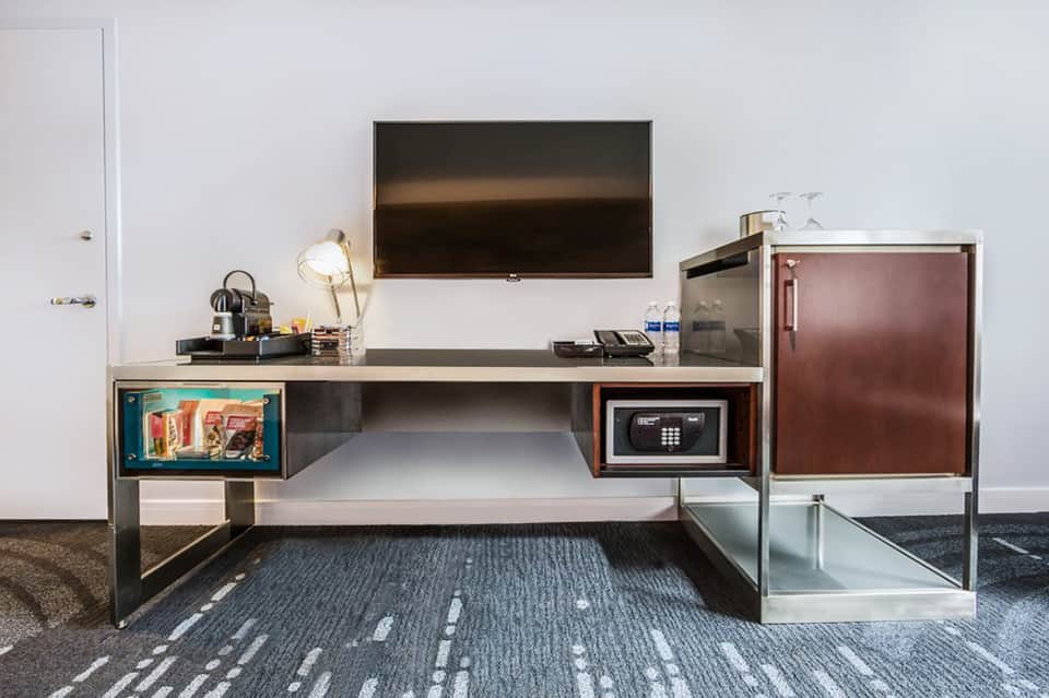 Modern hotel room desk with TV, safe, minibar, and work lamp on textured carpet flooring