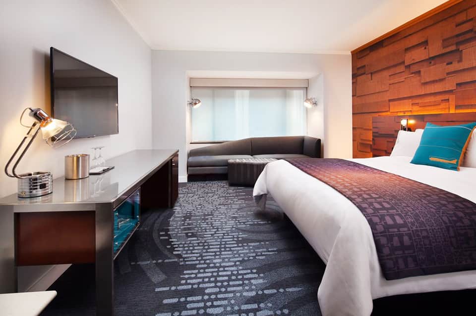 Modern hotel suite with king bed, work desk, flat-screen TV, and brick accent wall with seating area