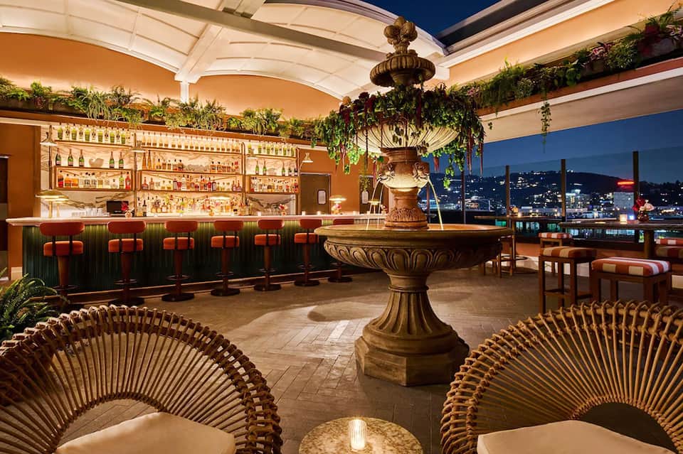 Upscale rooftop bar with golden fountain, woven chairs, city skyline views, and illuminated liquor shelves