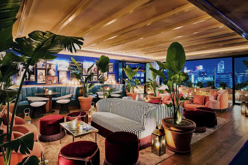 Luxury lounge with blue and coral seating, striped sofa, potted plants, wooden ceiling, and city skyline views at night