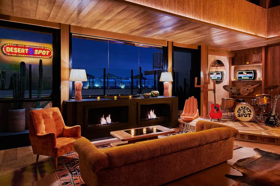 Retro-modern lounge with orange velvet seating, double-sided fireplace, desert neon sign, red guitar, and city skyline views