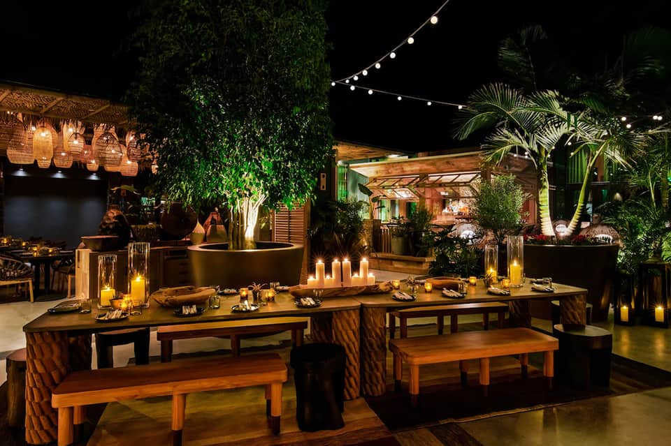 Outdoor evening event space with long wooden tables, benches, string lights, potted plants, and tropical landscaping