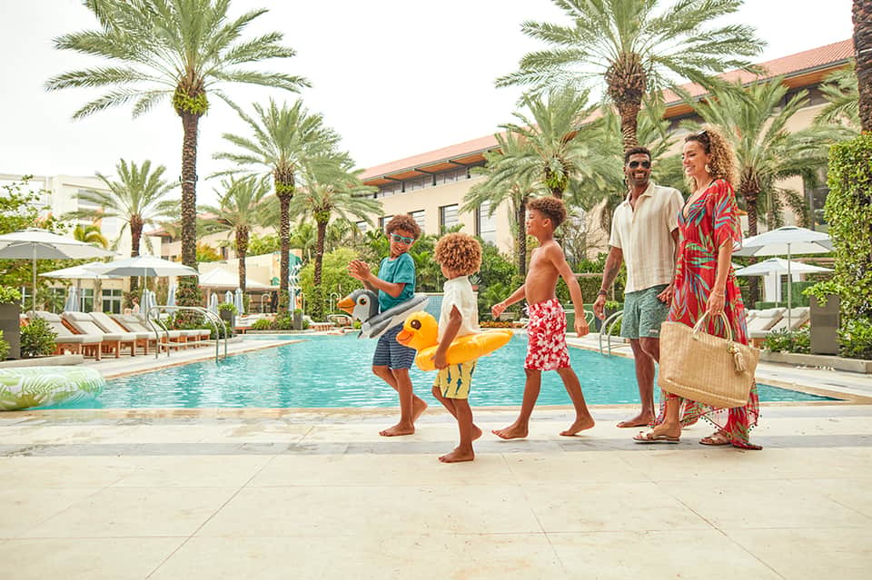 Family enjoying poolside with children holding duck floats near palm trees and resort buildings