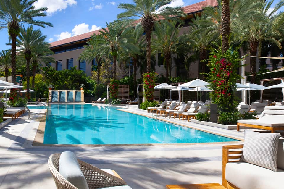 Luxury resort pool with palm trees, lounge chairs, white umbrellas, and Mediterranean-style building
