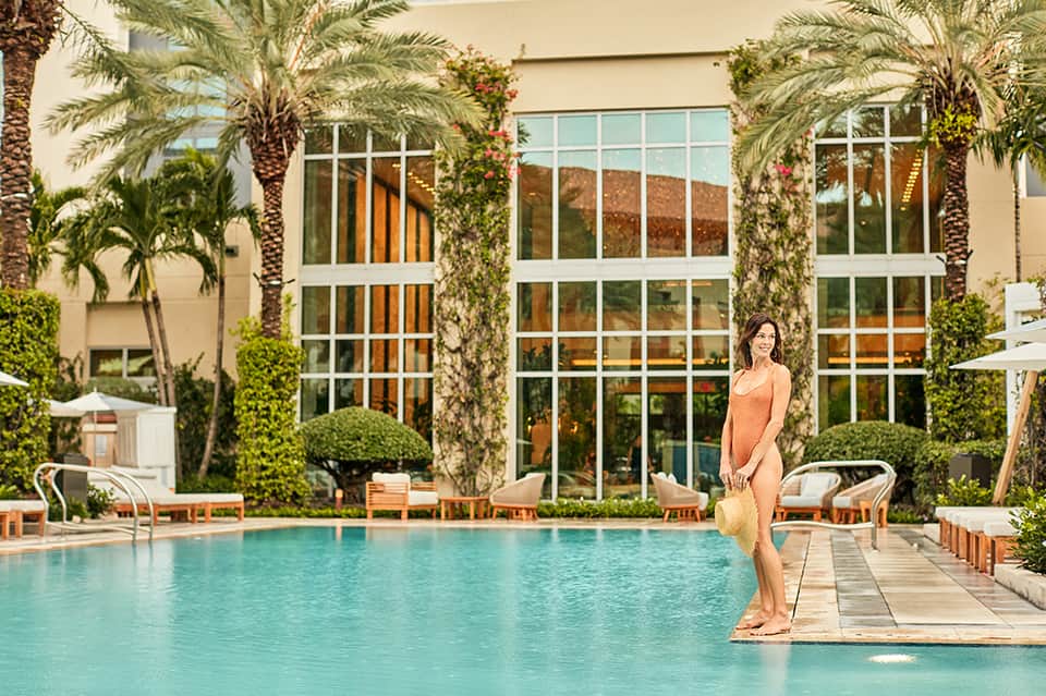 Woman in orange swimsuit standing on pool deck beside turquoise pool, flanked by palm trees and modern building