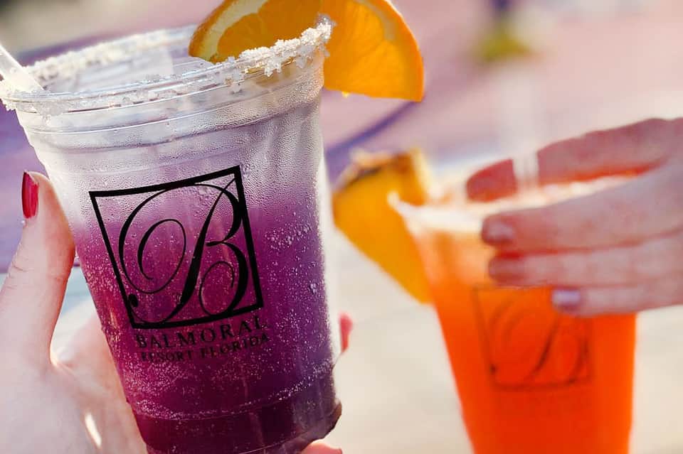 Purple-hued cocktail in branded glass with orange slice garnish, held in sunlight