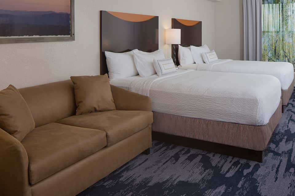 Modern hotel room with two queen beds, white bedding, tan sofa, and patterned carpet flooring