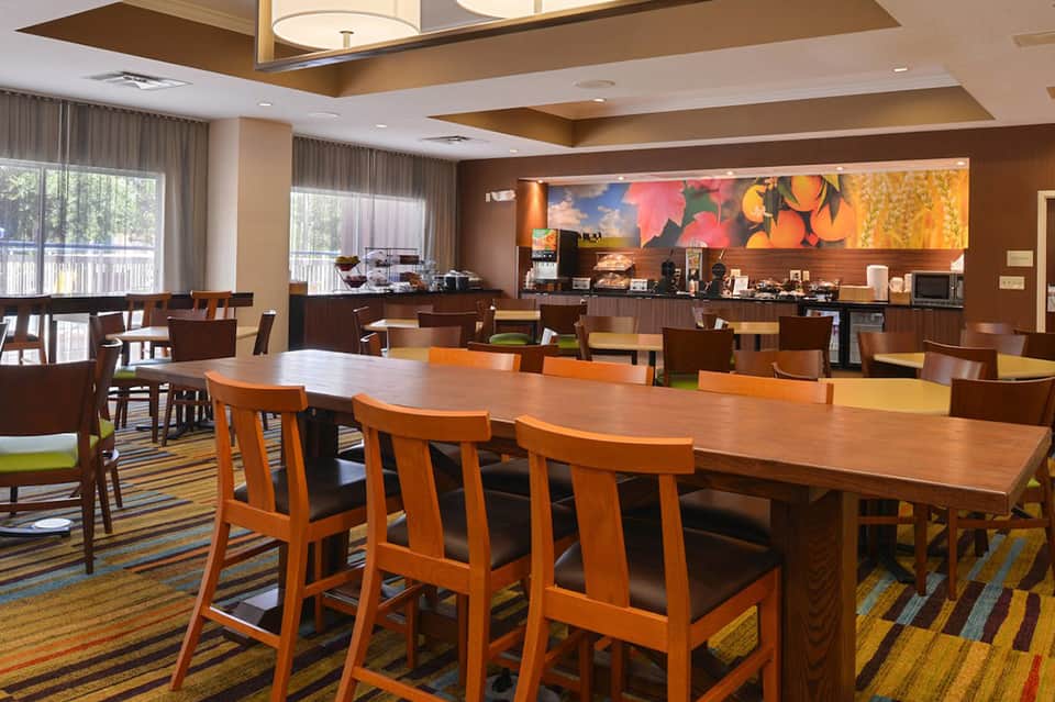 Modern hotel breakfast area with wooden dining tables, orange chairs, colorful fruit artwork, and floor-to-ceiling windows