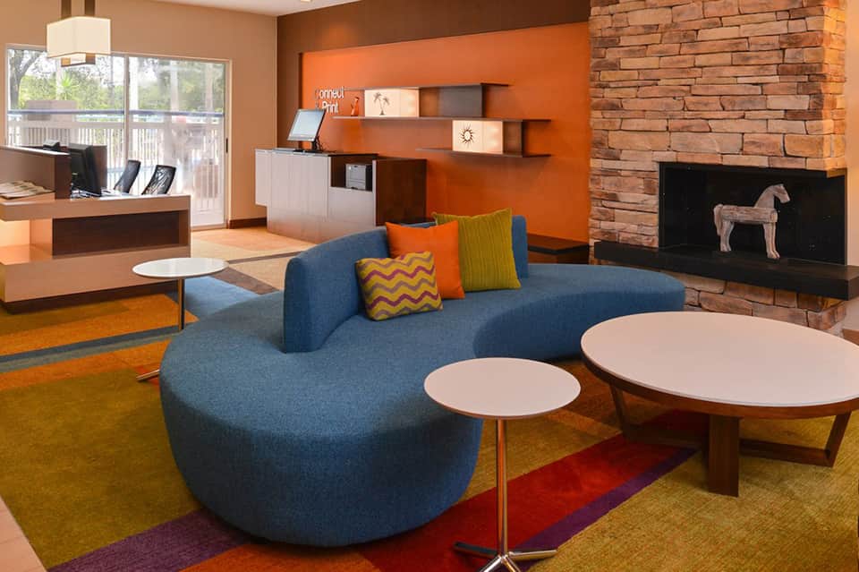Modern office lounge with curved blue sofa, colorful pillows, round tables, and orange accent wall with brick fireplace