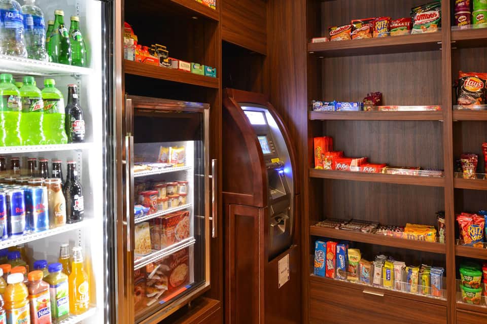 Hotel convenience store with refrigerated beverages, snacks, ATM, and wooden shelving displaying packaged goods