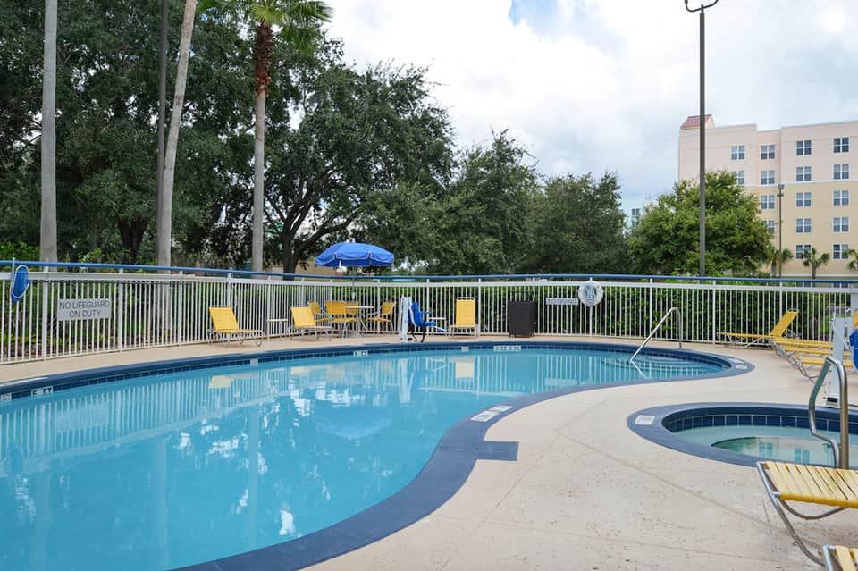 Resort swimming pool with blue water, yellow lounge chairs, white railing, palm trees, and a multi-story hotel building in background
