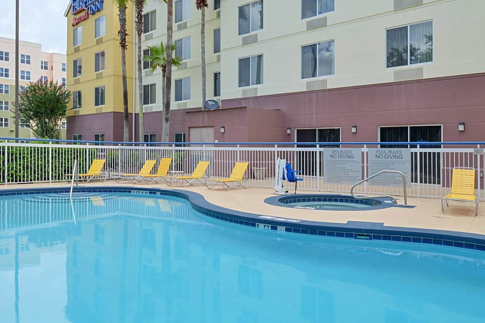 Outdoor swimming pool with yellow lounge chairs, hot tub, and hotel buildings in background