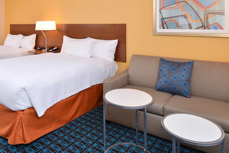 Modern hotel room with queen beds, orange bedding, tan sofa, blue patterned rug, and yellow walls
