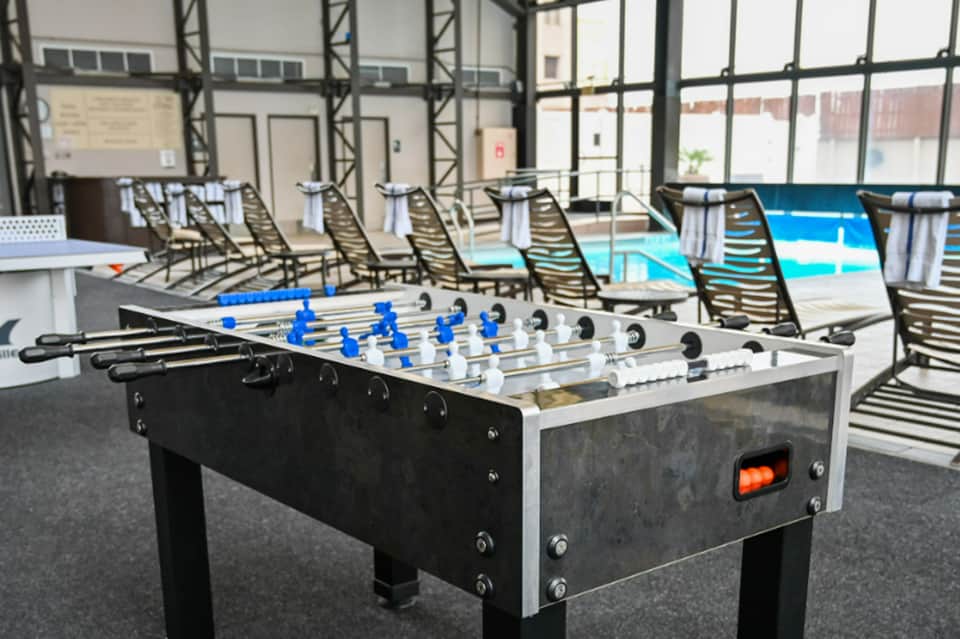 Foosball table by pool with lounge chairs and large windows overlooking waterfront