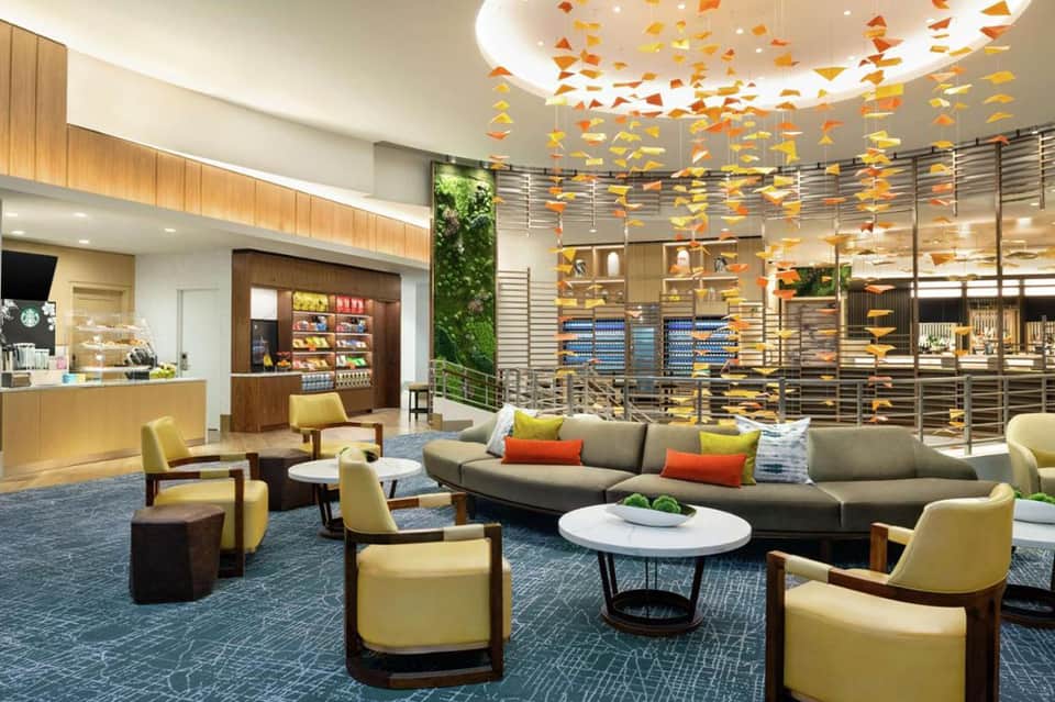 Modern hotel lobby with yellow armchairs, gray sofa, hanging orange decorations, and Starbucks counter