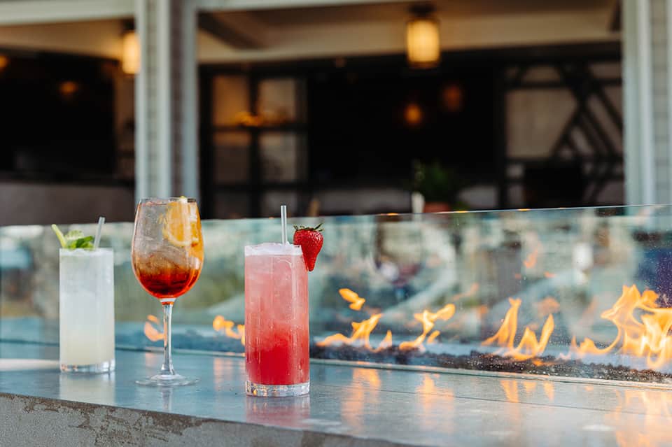 Three colorful cocktails on a fire pit bar counter with flames in background