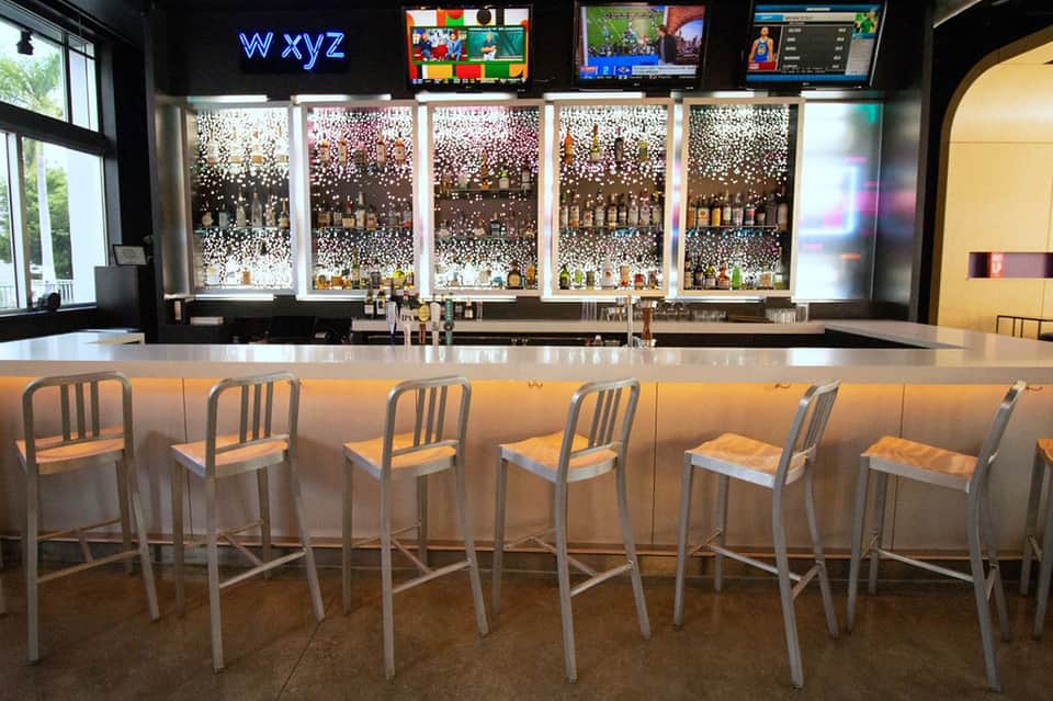 Modern bar with illuminated shelving displaying bottles, metal bar stools, and screens above the counter