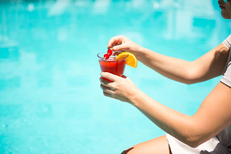 Person holding a red cocktail with orange slice garnish by a bright turquoise swimming pool