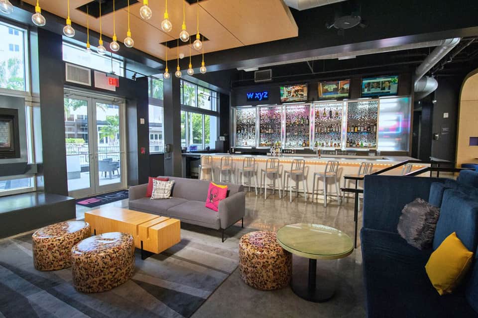 Modern hotel bar and lounge with pendant lighting, gray seating, patterned ottomans, and floor-to-ceiling windows