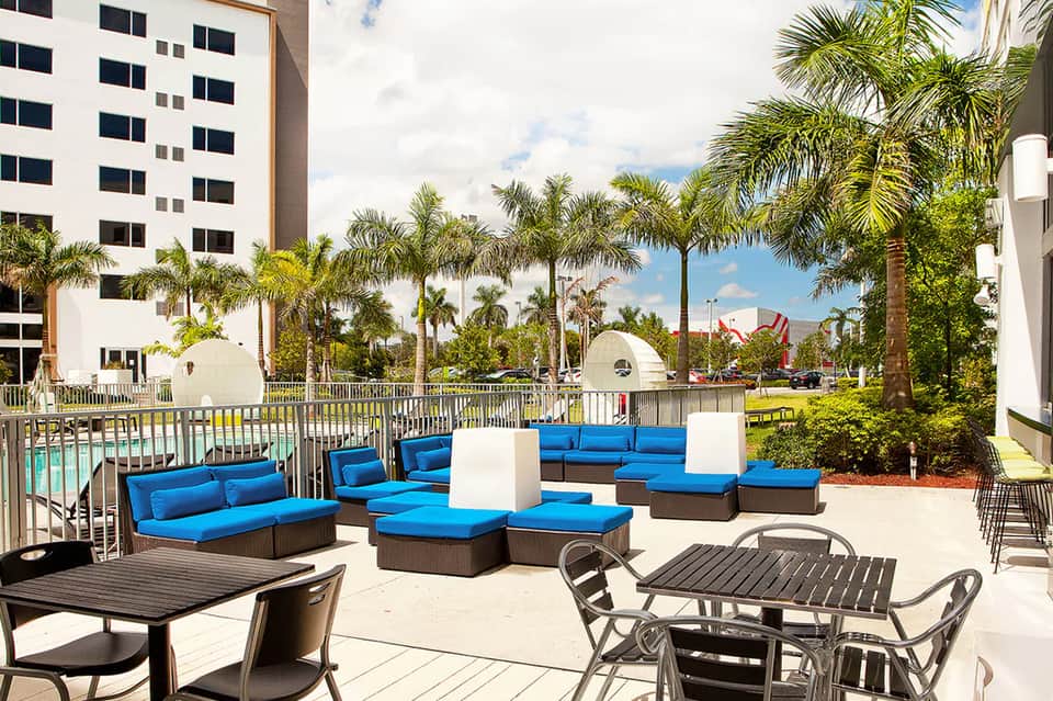 Resort poolside lounge area with blue cushioned seating, palm trees, and white building in background