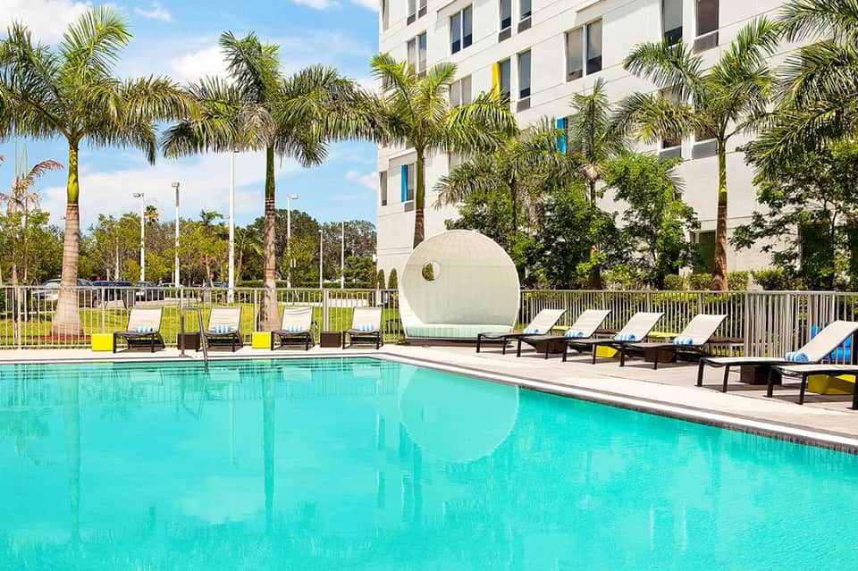 Resort pool with turquoise water, palm trees, white loungers, and modern building